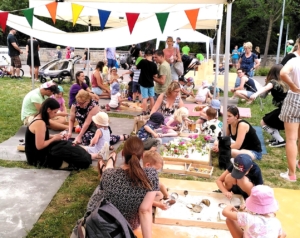 SpielOase Familienspielraum Viele Familien mit kleinen Kindern sitzen an flachen Holzkästen, in denen Spielmatrial liegt unter einem Pavillion im Freien
